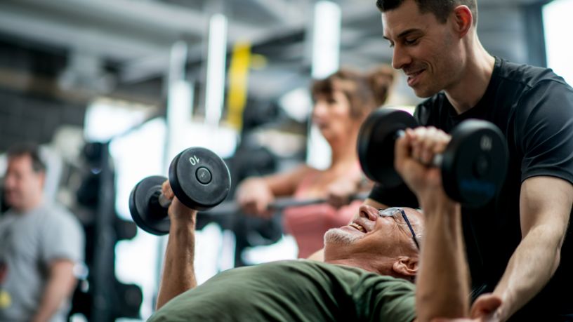 A trainer helps a man lift weights at a gym.