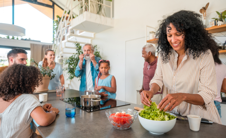 A woman cheerfully prepares a leafy salad in a bowl, while surrounded by family of all ages in a nice modern kitchen.