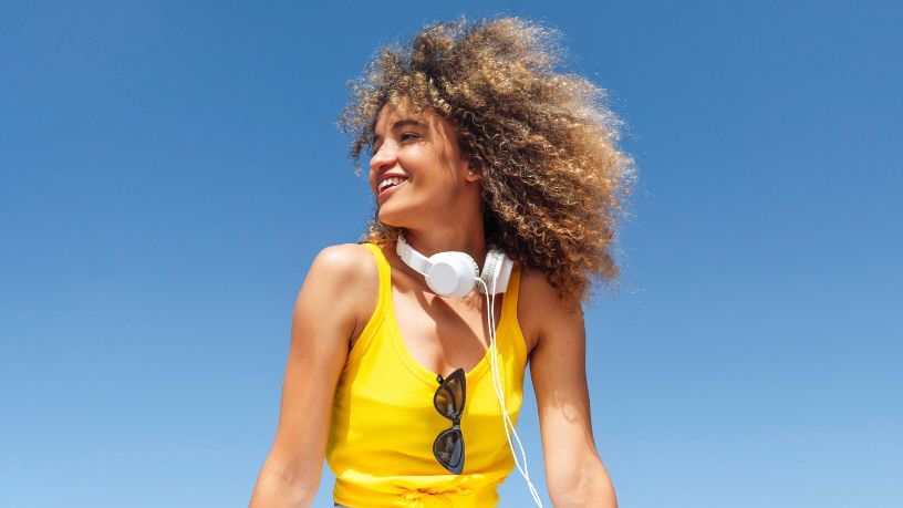 A woman with headphones and sunnies smiles in the sun.