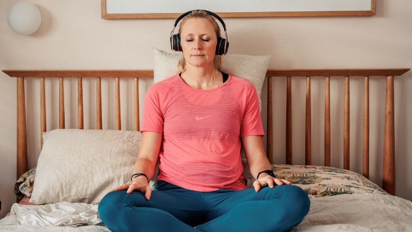 A person wearing headphones sits on their bed with their eyes closed, meditating.