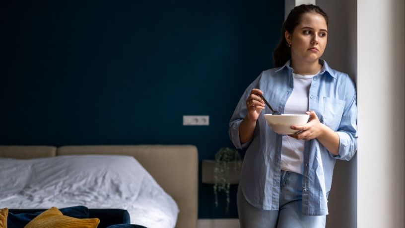 A woman leans against her bedroom wall while eating breakfast.