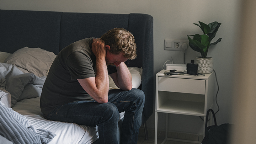A man sitting on his unmade bed with his head in his hands.