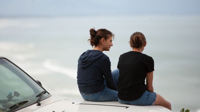 Two people sit on a car bonnet overlooking the water.