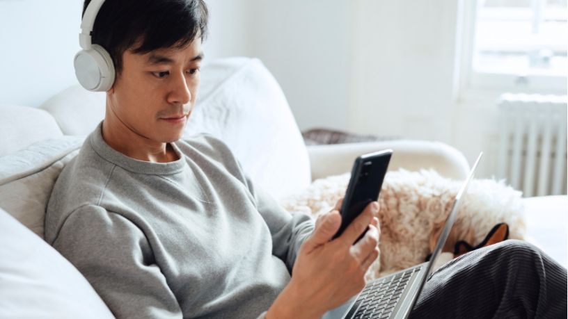 A man wearing headphones looks at his phone and balances his laptop on his legs.