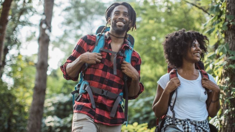 A man and a woman walk through nature with hiking packs on their backs.
