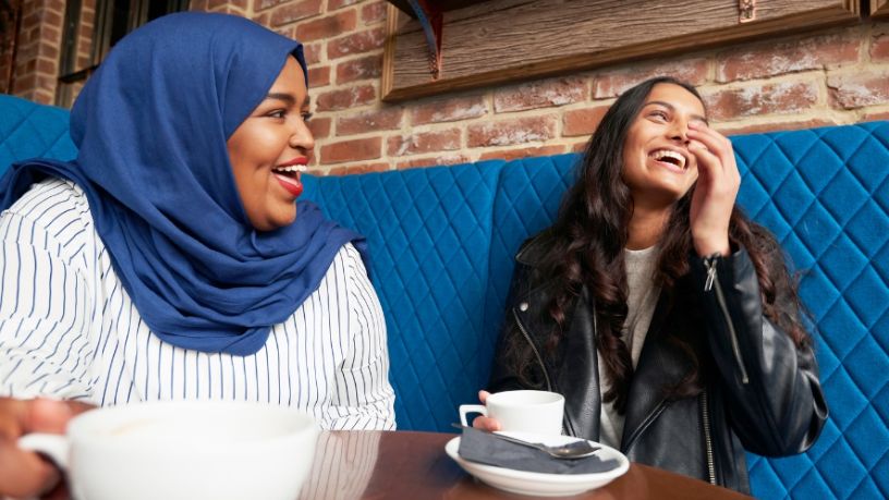 Two women sit in a café booth holding coffee cups and laughing.