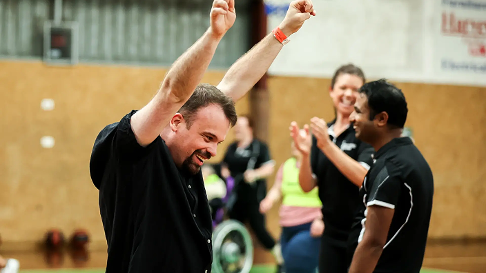 Indoor court setting. Adult man in foreground raising both arms in celebration. Other people clapping in background, including a person using a sports wheelchair.
