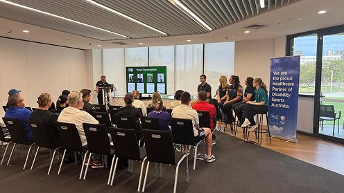 A group of people seated in a bright, modern community room attending a presentation about disability sports.