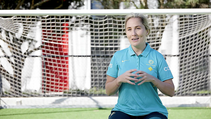 Rae smiling in front of a white soccer net, wearing a light blue Australian Paralympic team polo shirt.