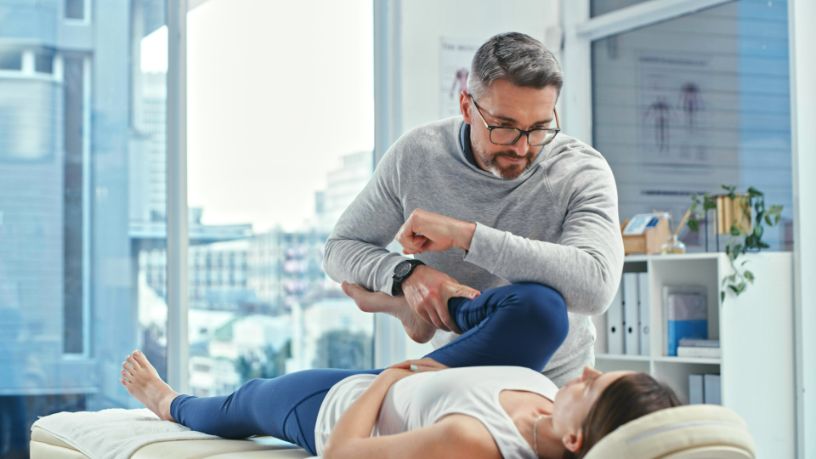 A woman lays on a treatment bed while a physiotherapist helps her stretch her leg and knee.
