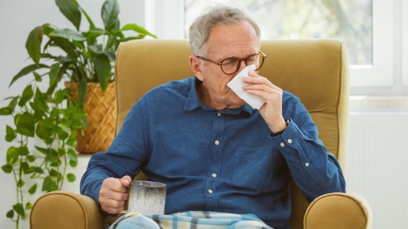 A man sits in an armchair, wiping his nose with a tissue and holding a mug.