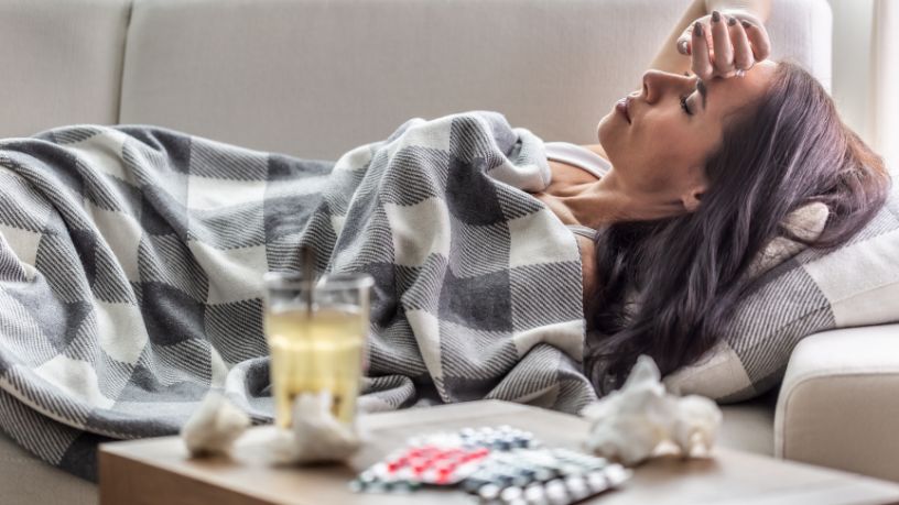 A woman laying on the couch with some cold and flu tablets at her side.