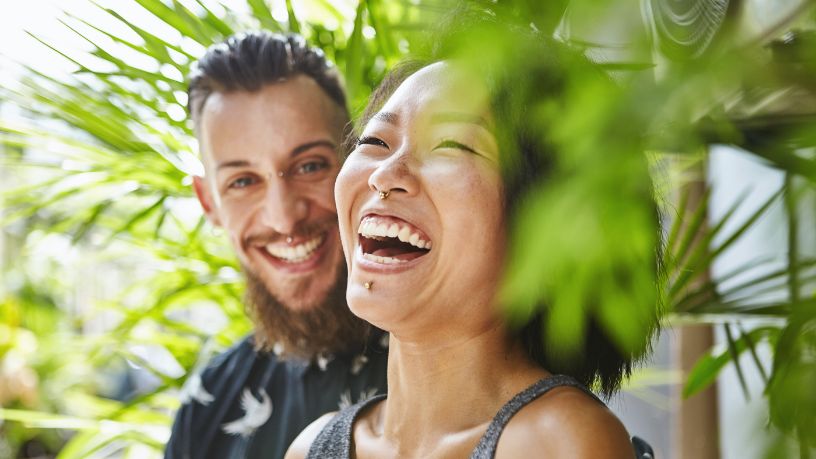 A man and woman laugh, standing between pot plants.