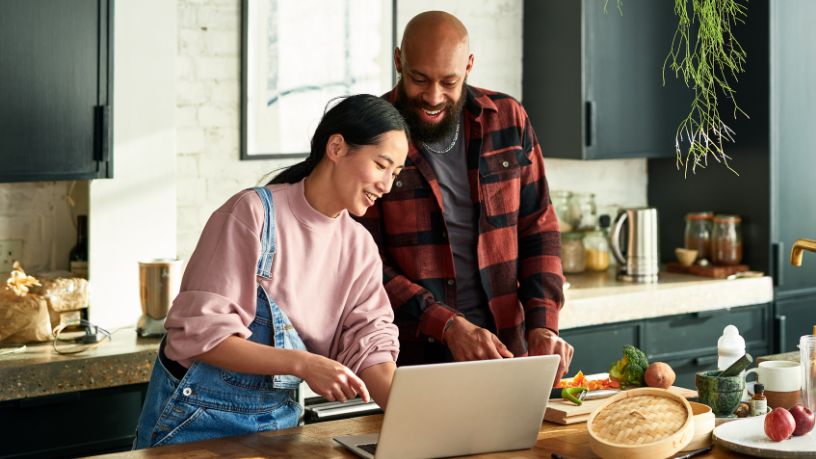 A man and woman consult their laptop while making a meal.