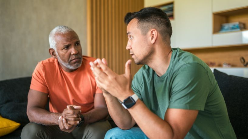 Two men have a serious conversation on a couch.