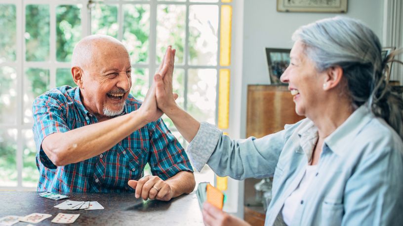 An older man and woman high-five while playing cards in their living room.