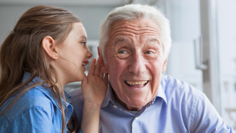 A young girl whispers in her grandad's ear as he laughs.