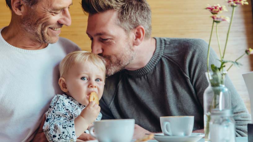 Two men sit with their baby while one kisses him on the head.