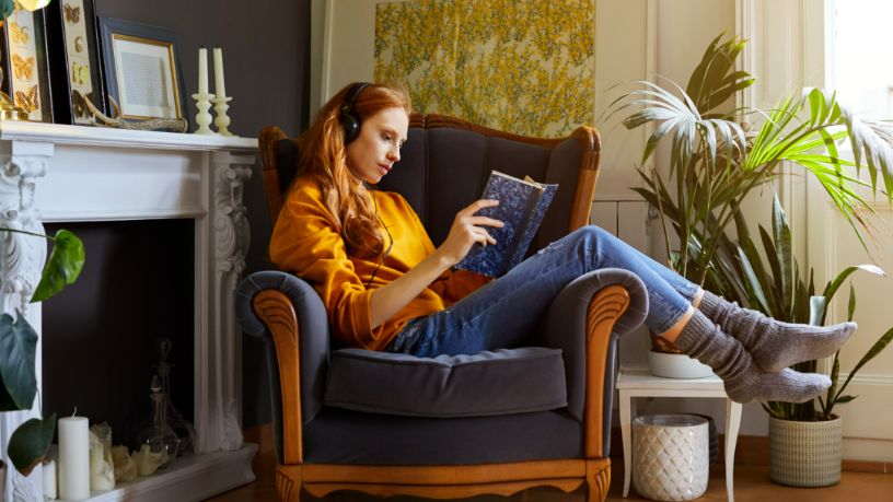 A woman reads a book while sitting sideways on an armchair.