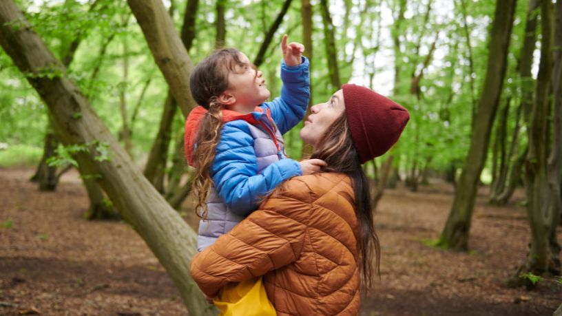 A woman holds her young daughter in the woods as she points to the sky.