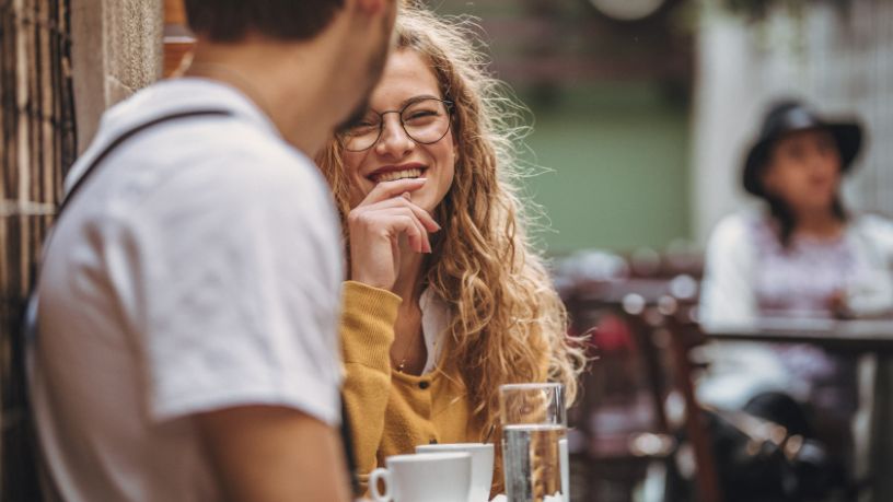A woman smiles as she drinks coffee outdoors with a man.