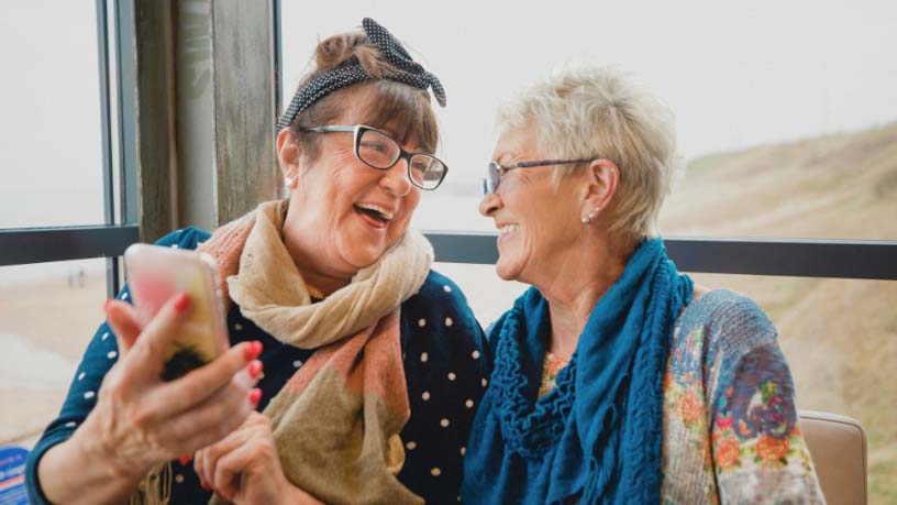 Two women site and laugh while looking at a phone.