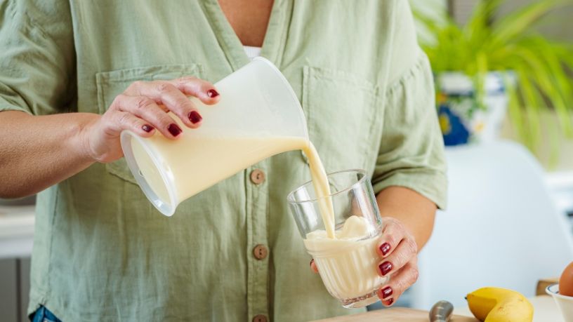 A person in a green shirt pours a banana smoothy from a jug into a glass.