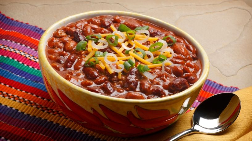 A bowl of black bean chilli rests on a table with a spoon next to it.