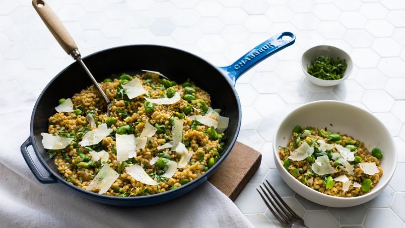 A cast iron pan with pearl barley risotto lays on a table with a bowl of the dish to its side.