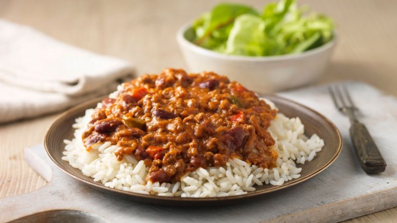 A plate of beef chilli on rice sits next to a side salad on a dining table.