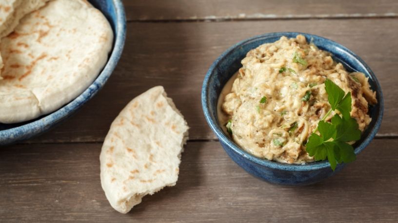 A torn off piece of pita bread lays on a table between a plate of pita and a bowl of baba ghanoush.