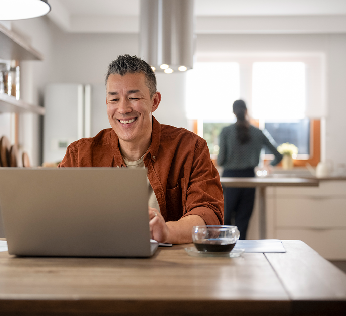 Man sits at a dining table with his laptop and a cup of coffee, while his partner works in the kitchen in the background.