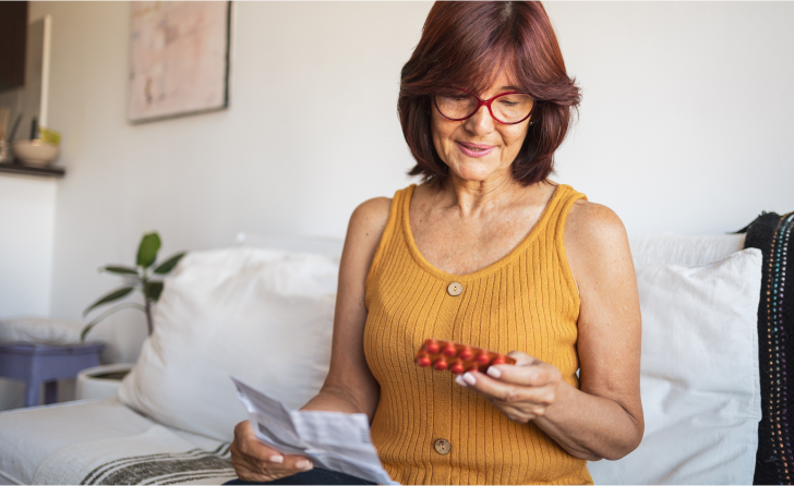 A middle-aged lady sits on her bed, looking at some red tablets she's holding in one hand.