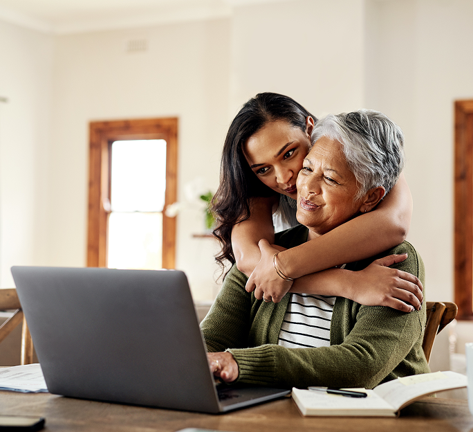 An older woman using a laptop is embraced by a younger woman, illustrating family members supporting each other in researching health options.