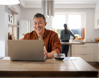 Man sits at a dining table with his laptop and a cup of coffee, while his partner works in the kitchen in the background.