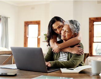 An older woman using a laptop is embraced by a younger woman, illustrating family members supporting each other in researching health options.