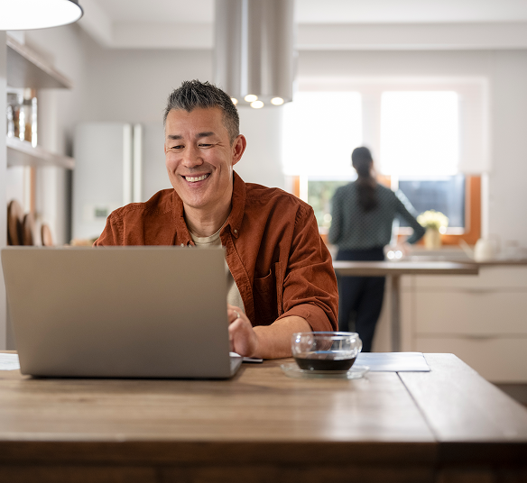 Man sits at a dining table with his laptop and a cup of coffee, while his partner works in the kitchen in the background.