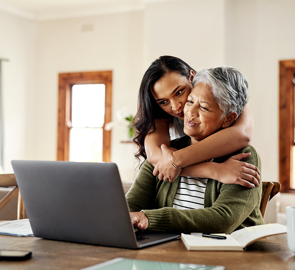 An older woman using a laptop is embraced by a younger woman, illustrating family members supporting each other in researching health options.